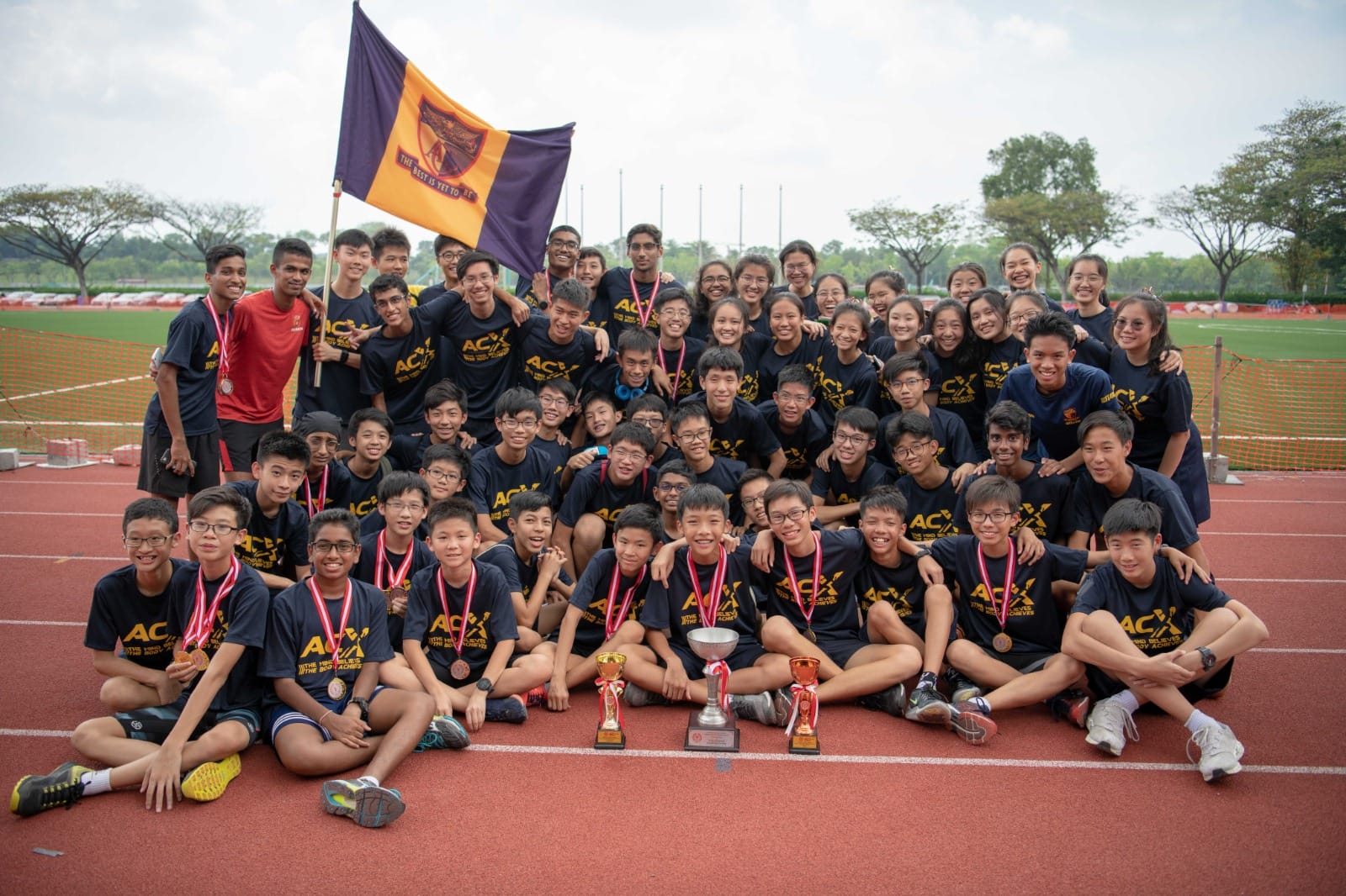 Large group of students in matching navy ACX shirts pose on a red track with trophies, medals, and a purple flag.